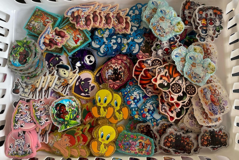 May include: A collection of assorted colourful character stickers featuring various cartoon figures, including Tweety Bird, dalmatians, and fantasy creatures, arranged in a white plastic bin with a grid pattern, showcasing diverse shapes and designs.