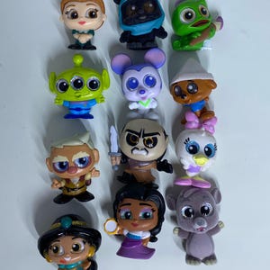 May include: A collection of twelve small, stylized plastic character figurines featuring various animated figures with large, glittery eyes. The set includes diverse characters with distinct outfits, colors, and accessories, arranged in a grid on a white surface.