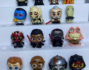 May include: A collection of sixteen small Star Wars character figurines arranged on a white tiered display stand. The figures feature stylised designs with large eyes and detailed outfits, including characters like Darth Vader, Kylo Ren, and Darth Maul.