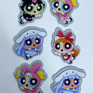 May include: Six glittery stickers featuring cartoon characters with large eyes. The designs include characters dressed in black with a skull, pink with bunny ears, and blue with long hair and white hoods, all with vibrant colors and expressive faces.
