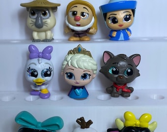 May include: A collection of twelve small, stylised character figurines featuring various animated figures with large, glittery eyes. The figures include human-like characters, animals, and creatures in diverse colours, arranged in four rows on a white stand.