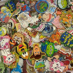 May include: A large collection of assorted colorful stickers featuring various cartoon characters, including Disney, Pixar, and classic animation figures. The stickers have diverse shapes, glittery textures, and vibrant printed designs.
