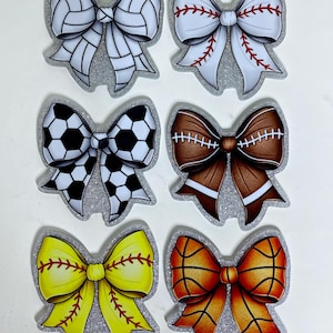 May include: Six decorative hair bows with a silver glitter border, each featuring a different sports ball pattern: a white volleyball, a white baseball with red stitching, a black and white soccer ball, a brown football, a yellow softball, and a basketball.