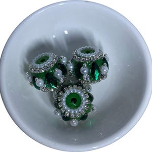 May include: Three round beads featuring a vibrant green base, decorated with small white pearls and clear rhinestones arranged in a circular pattern around the center hole, creating a textured and reflective surface on each individual bead.