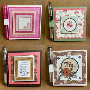 May include: Four handmade square notepads with pens. Each notepad features layered paper designs in pink, green, brown, and purple. Some have floral embroidery hoop designs or inspirational quotes. Each notepad has a pen attached.