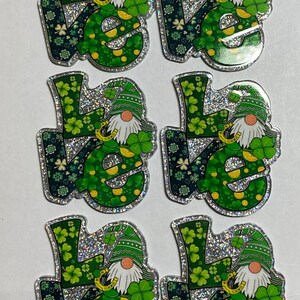 May include: Six St. Patrick's Day themed embellishments. Each features the word "Love" in green with shamrocks and a gnome wearing a green hat. The edges are outlined with silver glitter.