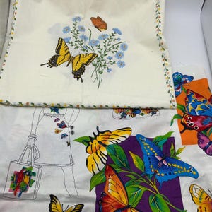 May include: A white fabric panel featuring a large yellow and black butterfly, a smaller orange butterfly, and blue flowers. Below, a second fabric piece displays a line drawing of a figure carrying a tote bag, alongside colorful butterflies.