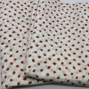 May include: A folded piece of fabric featuring an off-white background with a repeating pattern of small red strawberries with tiny green leaves. The textile has a soft, lightweight appearance with a consistent print distributed across the surface.