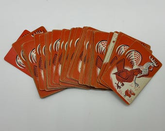 Puede incluir: Una baraja de cartas rectangulares dispuestas en abanico, cada una con un fondo naranja vibrante y una ilustración estilizada de un gallo y dos polluelos en tinta blanca, negra y roja, mostrando un diseño gráfico consistente de temática rural.