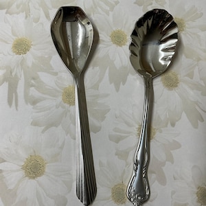 May include: Two silver-toned metal spoons with polished finishes. The left spoon features a hexagonal bowl and a handle with vertical fluted lines. The right spoon has a scalloped, shell-shaped bowl and a handle with embossed floral detailing.