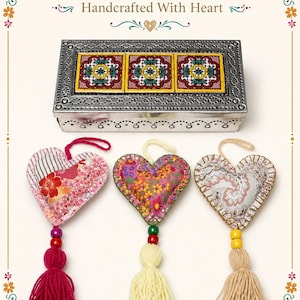 Puede incluir: Una caja rectangular de metal plateado con tres azulejos florales incrustados, situada sobre tres corazones de tela rellenos. Cada corazón presenta un tejido estampado único, un lazo para colgar, una cuenta de color y una borla de lana.