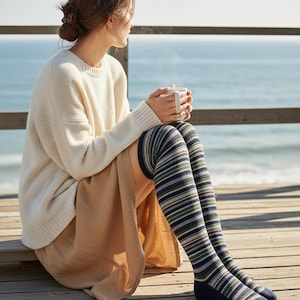May include: Thigh-high socks featuring a horizontal striped pattern in shades of navy blue, olive green, light grey, and white. The socks have a solid navy blue toe and heel section, extending above the knee with a ribbed texture throughout the knit fabric.