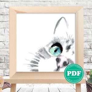 May include: A black and white cross stitch pattern of a cat's face with one blue eye looking to the right. The pattern is framed in a light wood frame and is sitting on a wooden easel.