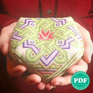 May include: A green, purple, and red cross stitch pattern with a geometric design. The pattern is stitched on a white fabric and is shaped like a cube. The cube is being held in the hands of a person.