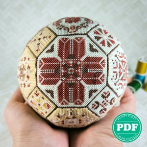 May include: A cross stitch ball ornament with a geometric pattern in shades of brown, cream, and red. The ball is held in two hands.