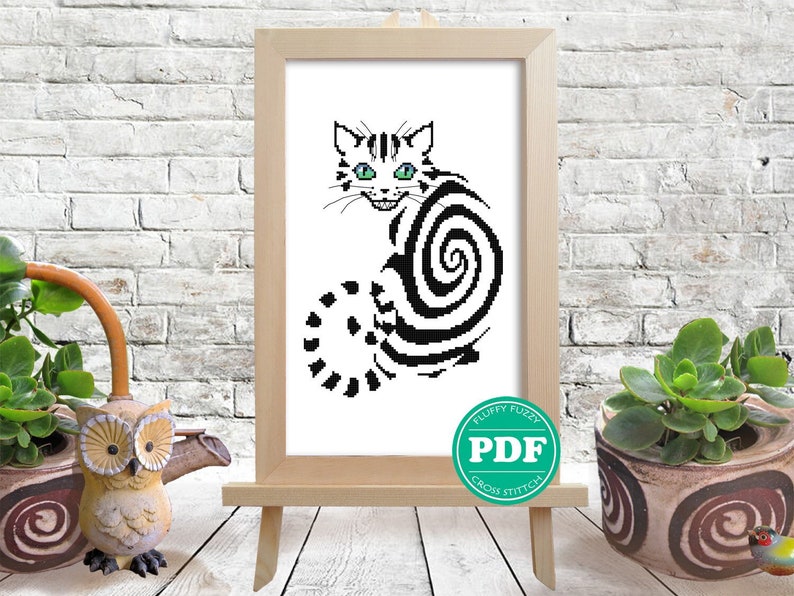 May include: Black and white cross stitch pattern of a cat curled up in a spiral shape. The cat has green eyes and a mischievous grin. The pattern is framed and displayed on an easel.