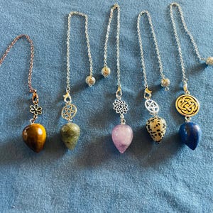 May include: Collection of dowsing pendulums with various gemstone pendants. The pendulums feature different chain styles and decorative charms, including a flower, star, and Celtic knot. Gemstones include tiger's eye, amethyst, and lapis lazuli.