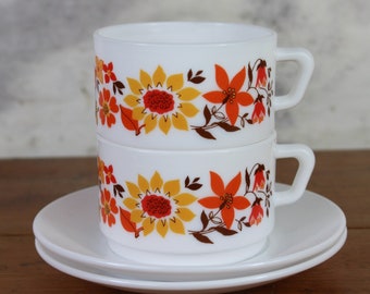 May include: Two white ceramic mugs with a floral pattern in shades of orange, yellow, and brown. The mugs are stacked on top of each other and are sitting on a white saucer.
