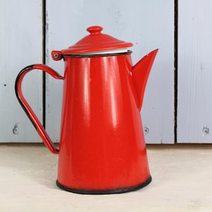 Può includere: Caffettiera in smalto rosso con coperchio bianco e manico nero. La caffettiera è appoggiata su una superficie bianca.