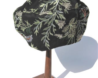 May include: A black hat with intricate floral embroidery in shades of green, gold, and white. The hat is displayed on a wooden hat stand, showcasing its detailed design and craftsmanship. The hat's fabric appears to have a soft texture.