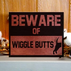 May include: A wooden sign with the words "BEWARE OF WIGGLE BUTTS" in a bold, rustic font. The sign has a black background with a reddish-brown border and text. A silhouette of a dog's rear end is on the right side.