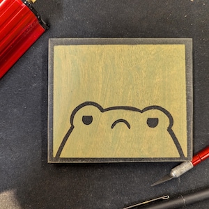 May include: A square wooden block with a cartoon frog face etched into it. The frog has a grumpy expression with a curved mouth and two oval eyes. The block is light brown with a dark brown border. A red pen and a black pen are in the background.