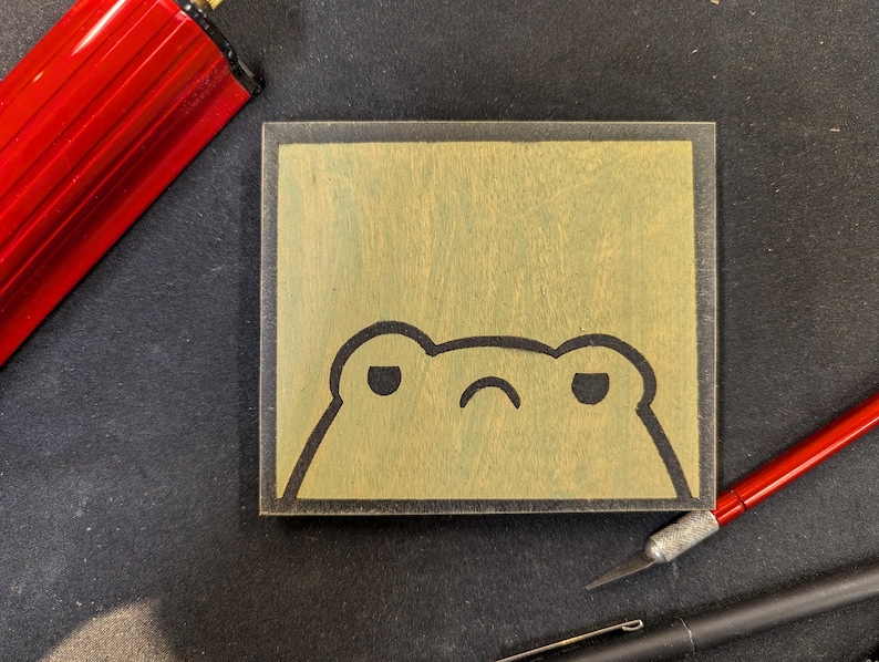 May include: A square wooden block with a cartoon frog face etched into it. The frog has a grumpy expression with a curved mouth and two oval eyes. The block is light brown with a dark brown border. A red pen and a black pen are in the background.