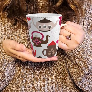 Puede incluir: Taza de cerámica blanca con un patrón repetido de teteras en tonos marrón, rosa y rojo. La taza tiene un asa curva. El diseño incluye corazones y detalles florales, creando una estética caprichosa con temática de té. La taza es sostenida por una persona.