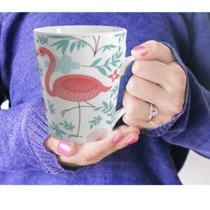 Pode incluir: Caneca de cerâmica branca com um design de flamingo rosa e detalhes florais. O flamingo tem um corpo texturizado e está sobre uma perna. A caneca tem uma alça curva e um acabamento liso e brilhante.