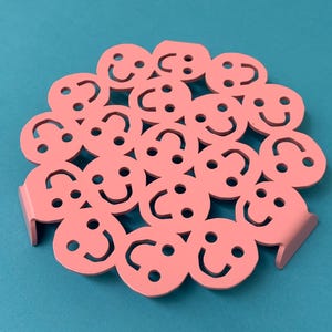 Puede incluir: Un salvamanteles circular de metal rosa con un patrón repetitivo de caras sonrientes troqueladas. El diseño consta de múltiples formas circulares interconectadas, cada una con dos ojos redondos y una boca curva, sobre pequeños pies metálicos.
