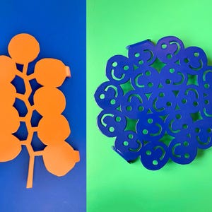 Puede incluir: Dos salvamanteles de metal, uno naranja y otro azul. El salvamanteles naranja tiene forma de rama con hojas redondas. El salvamanteles azul tiene forma de círculo con un patrón de caras sonrientes.