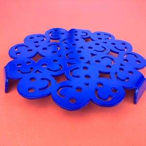 Op de afbeelding: Een blauwe metalen onderzetter met een cirkelvormig ontwerp met meerdere smiley-gezichten. De onderzetter heeft een verhoogde rand en twee kleine lipjes aan de zijkant.