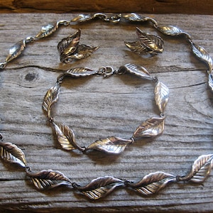 Può includere: Un set di collana e orecchini in argento con ciondoli a forma di foglia. La collana è realizzata con una catena di foglie interconnesse, mentre gli orecchini sono semplici motivi a foglia.