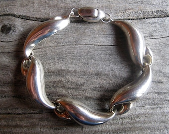 May include: A silver bracelet with a wavy, organic design. The bracelet is made of smooth, polished metal and has a clasp closure.