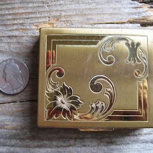 May include: A vintage gold-coloured compact with floral and scroll detailing. The square compact has a decorative border. A coin is next to it for size comparison. The compact is likely made of metal.