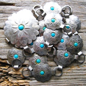 May include: A collection of silver-toned metal belt buckles with turquoise-colored centers. The buckles have a floral design and are connected by metal rings. The image is taken on a weathered wooden surface.