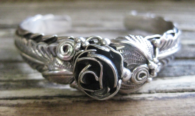May include: A silver bracelet with a black rose design. The bracelet is intricately detailed with swirls and leaves.