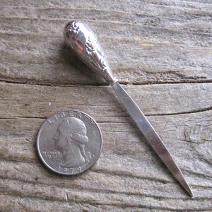 May include: A silver letter opener with a decorative floral design on the handle. The opener is next to a U.S. quarter coin from 1979. The opener is approximately 15 cm long.