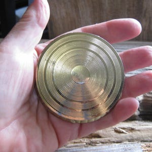 May include: A circular, gold-toned compact mirror with a pattern of concentric circles. The compact is held in a hand, highlighting its metallic finish and detailed surface. The mirror's design includes a central depression surrounded by textured rings.