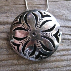May include: A silver-colored pendant necklace with a circular, openwork design. The pendant features a central hole and radiating, curved shapes with intricate cut-out details. The necklace hangs on a thin, silver chain.