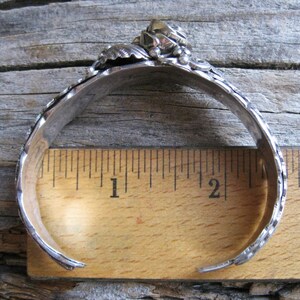 May include: A silver bracelet with a floral design and a single stone in the center. The bracelet is measured against a ruler, showing it is approximately 2 inches in diameter.