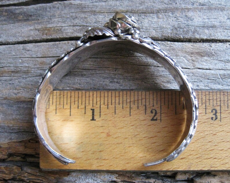 May include: A silver bracelet with a floral design and a single stone in the center. The bracelet is measured against a ruler, showing it is approximately 2 inches in diameter.