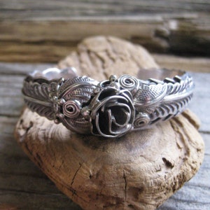 May include: A silver bracelet with a rose design. The rose is made of twisted wire and is surrounded by leaves. The bracelet is made of sterling silver and is a beautiful piece of jewelry.