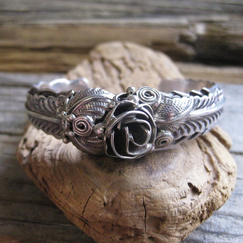 May include: A silver bracelet with a rose design. The rose is made of twisted wire and is surrounded by leaves. The bracelet is made of sterling silver and is a beautiful piece of jewelry.