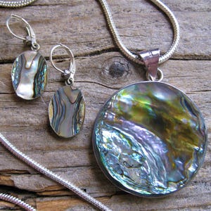 May include: A silver chain necklace with a large round abalone shell pendant. The pendant has iridescent green, blue, and purple colors. Matching earrings with smaller abalone shell drops are also included.