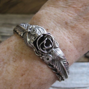 May include: A silver bracelet with a black rose design. The rose is made of metal and has a detailed design. The bracelet is made of silver and has a delicate design.