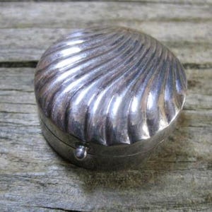 May include: A small, round, silver-colored metal pill box with a hinged lid. The top of the lid features a raised, swirling, shell-like pattern with deep grooves radiating from the center to the edge. A tiny, spherical metal knob is attached to the front.