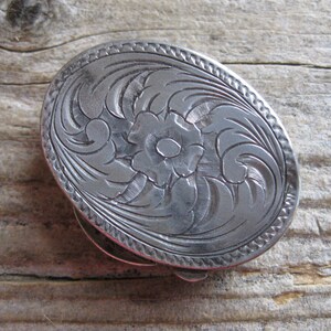 Puede incluir: Una hebilla de cinturón ovalada de plata con un intrincado diseño floral.