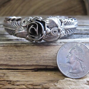 May include: A silver bracelet with a detailed rose design. The bracelet is shown next to a US quarter for size comparison.