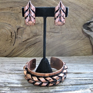 Könnte beinhalten: Ein Set aus Kupferohrringen und einem Armband. Die Ohrringe sind blattförmig und haben ein geometrisches Design. Das Armband besteht aus gedrehtem Kupferdraht und hat ein ähnliches Blattdesign.